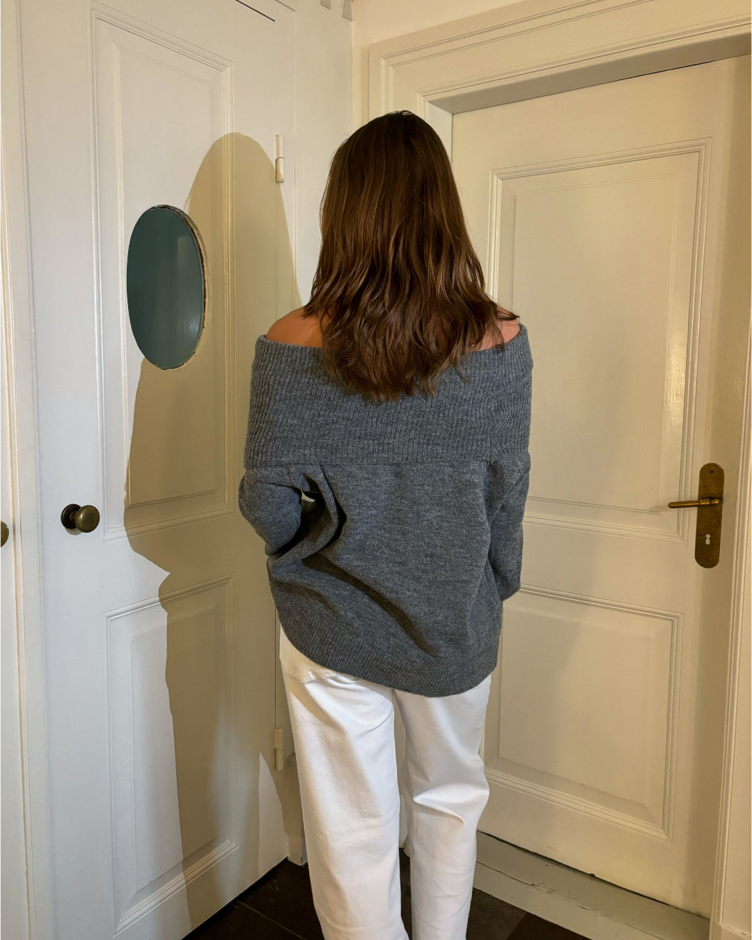 En person i en grå off-shoulder sweater og hvide bukser står med ryggen til i et rum med lyse døre og brunt gulv.