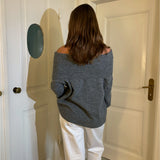 En person i en grå off-shoulder sweater og hvide bukser står med ryggen til i et rum med lyse døre og brunt gulv.