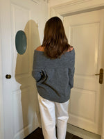 En person i en grå off-shoulder sweater og hvide bukser står med ryggen til i et rum med lyse døre og brunt gulv.
