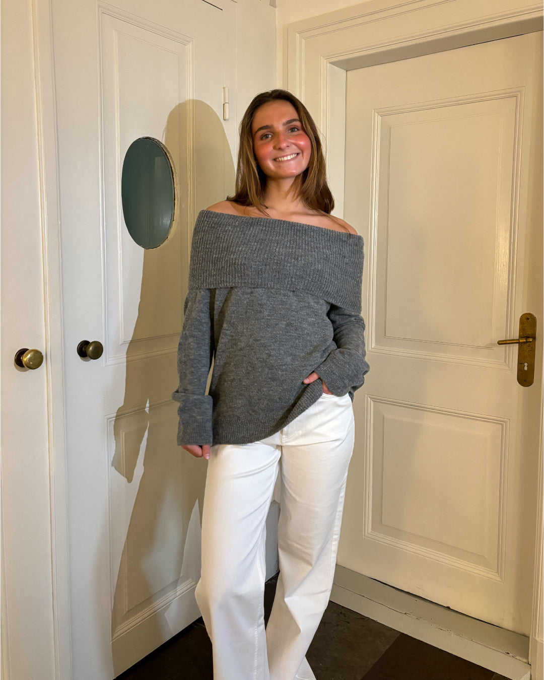 En person smiler iført en grå off-shoulder sweater og hvide bukser, står foran hvide døre med messinghåndtag.