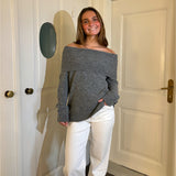 En person smiler iført en grå off-shoulder sweater og hvide bukser, står foran hvide døre med messinghåndtag.