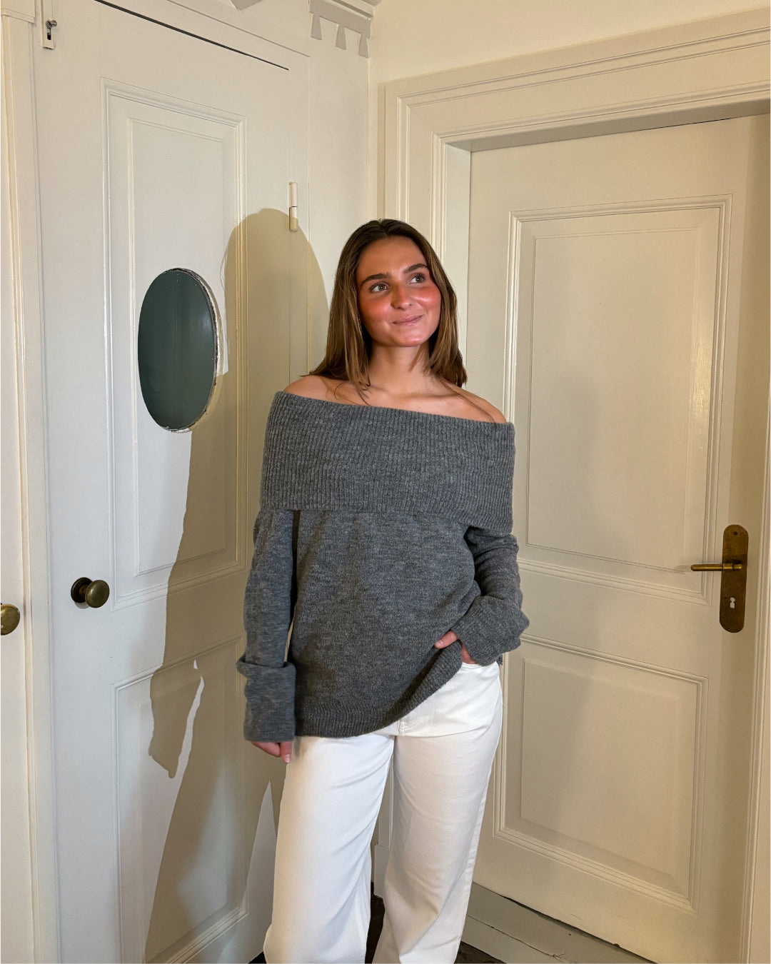En person iført en grå, skulderfri sweater og hvide bukser står smilende i et lyst rum med to hvide døre.