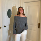 En person iført en grå, skulderfri sweater og hvide bukser står smilende i et lyst rum med to hvide døre.