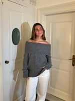 En person iført en grå, skulderfri sweater og hvide bukser står smilende i et lyst rum med to hvide døre.