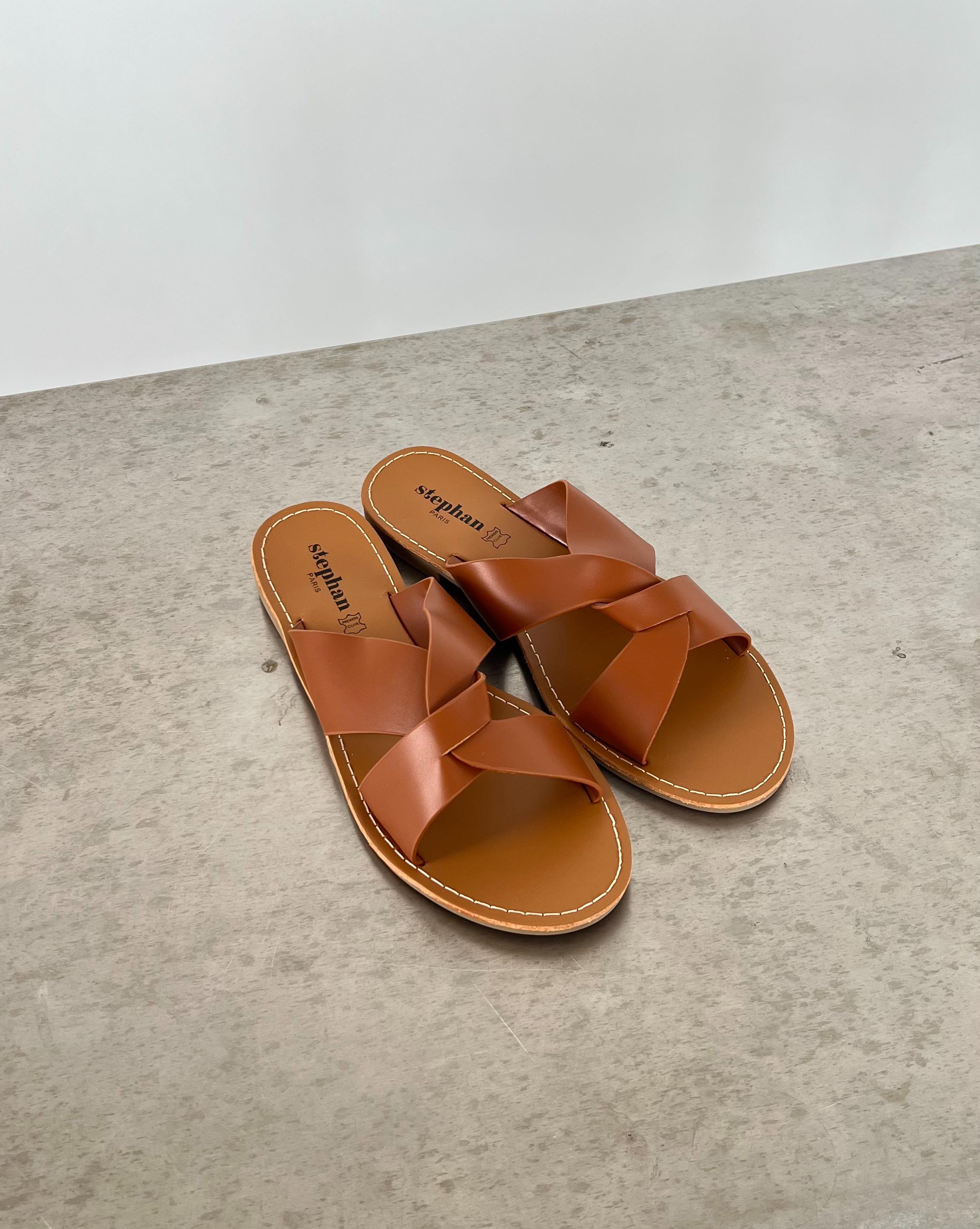 Brune sandaler med krydsede stropper ligger på en grå betonoverflade. Indersålen har teksten "stephan 87" trykt på.
