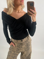 En person iført sort off-shoulder bluse og beige slangebukser tager en selfie med en telefon. Personen bærer en gylden halskæde, ring og har hånden i lommen.
