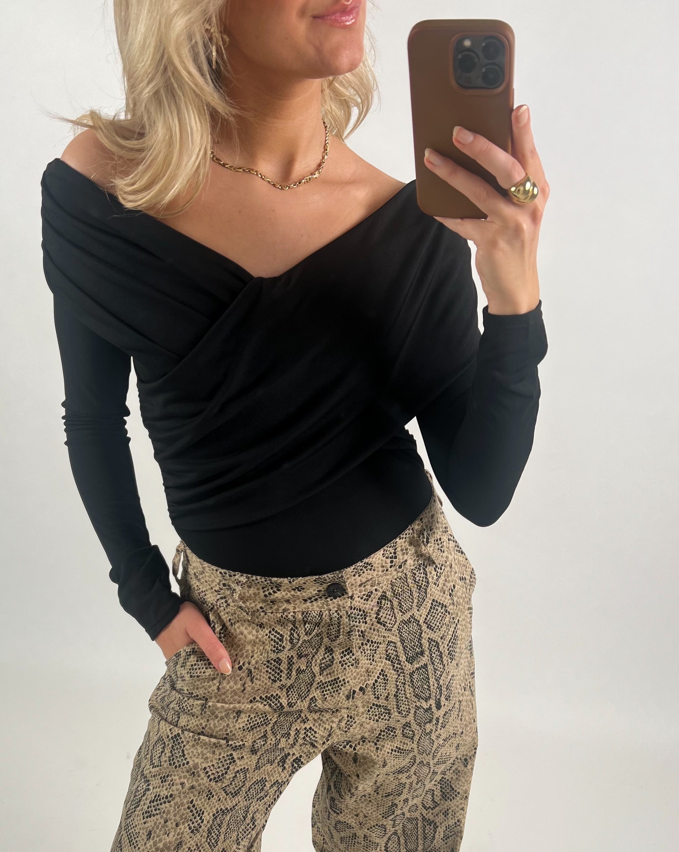 En person iført sort off-shoulder bluse og beige slangebukser tager en selfie med en telefon. Personen bærer en gylden halskæde, ring og har hånden i lommen.