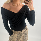 En person iført en sort off-shoulder bluse tager en selfie med en smartphone. De har blonde krøller, bærer en guldhalskæde og en ring, og har bukser med slangeskindsprint.