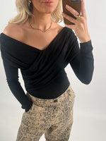 En person iført en sort off-shoulder bluse tager en selfie med en smartphone. De har blonde krøller, bærer en guldhalskæde og en ring, og har bukser med slangeskindsprint.