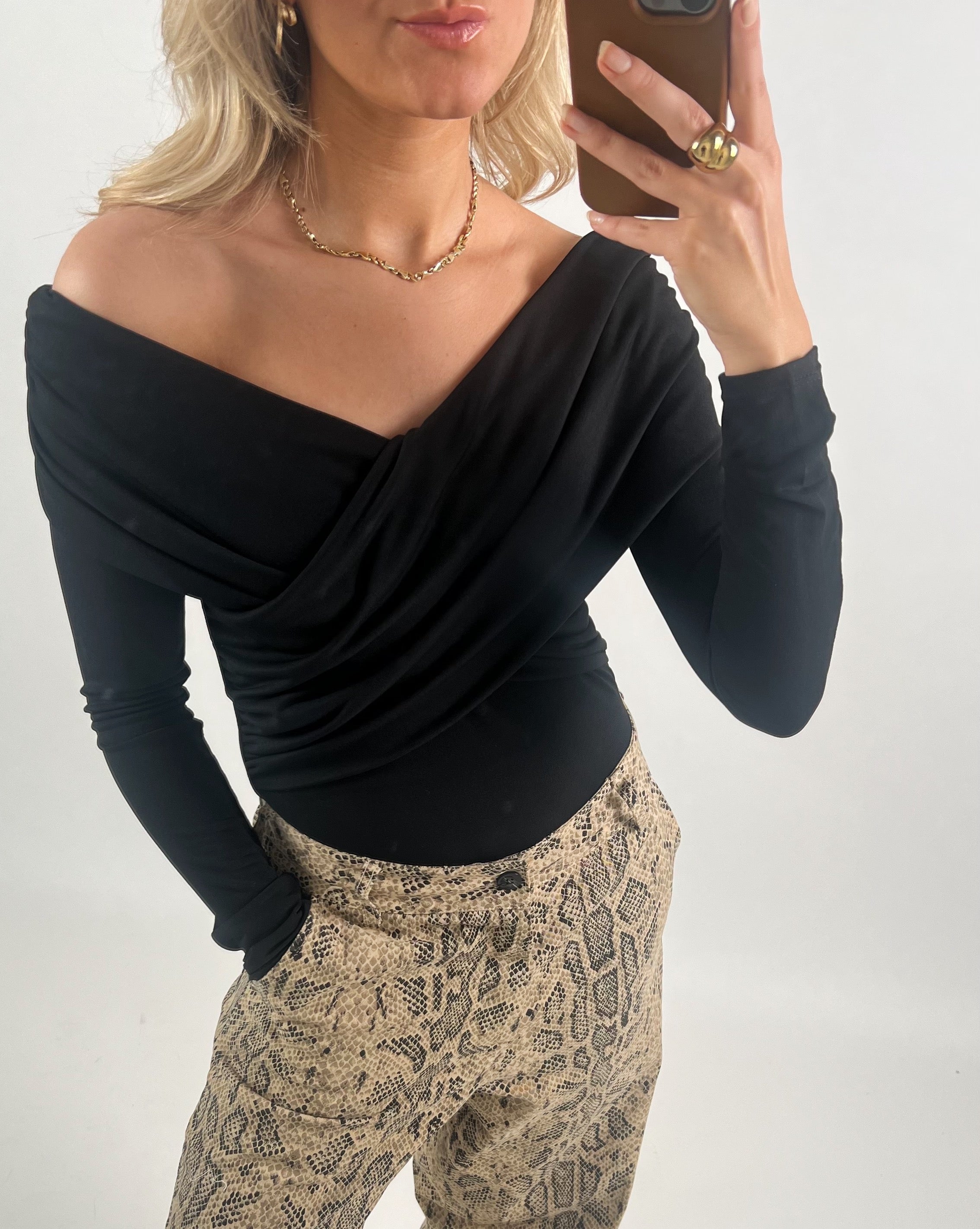 En person iført en sort off-shoulder bluse tager en selfie med en smartphone. De har blonde krøller, bærer en guldhalskæde og en ring, og har bukser med slangeskindsprint.