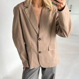 En person iført en beige blazer og grå bukser tager en selfie i et lyst rum. Personen holder en grøn mobiltelefon nær ansigtet.