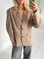 En person iført en beige blazer og grå bukser tager en selfie i et lyst rum. Personen holder en grøn mobiltelefon nær ansigtet.