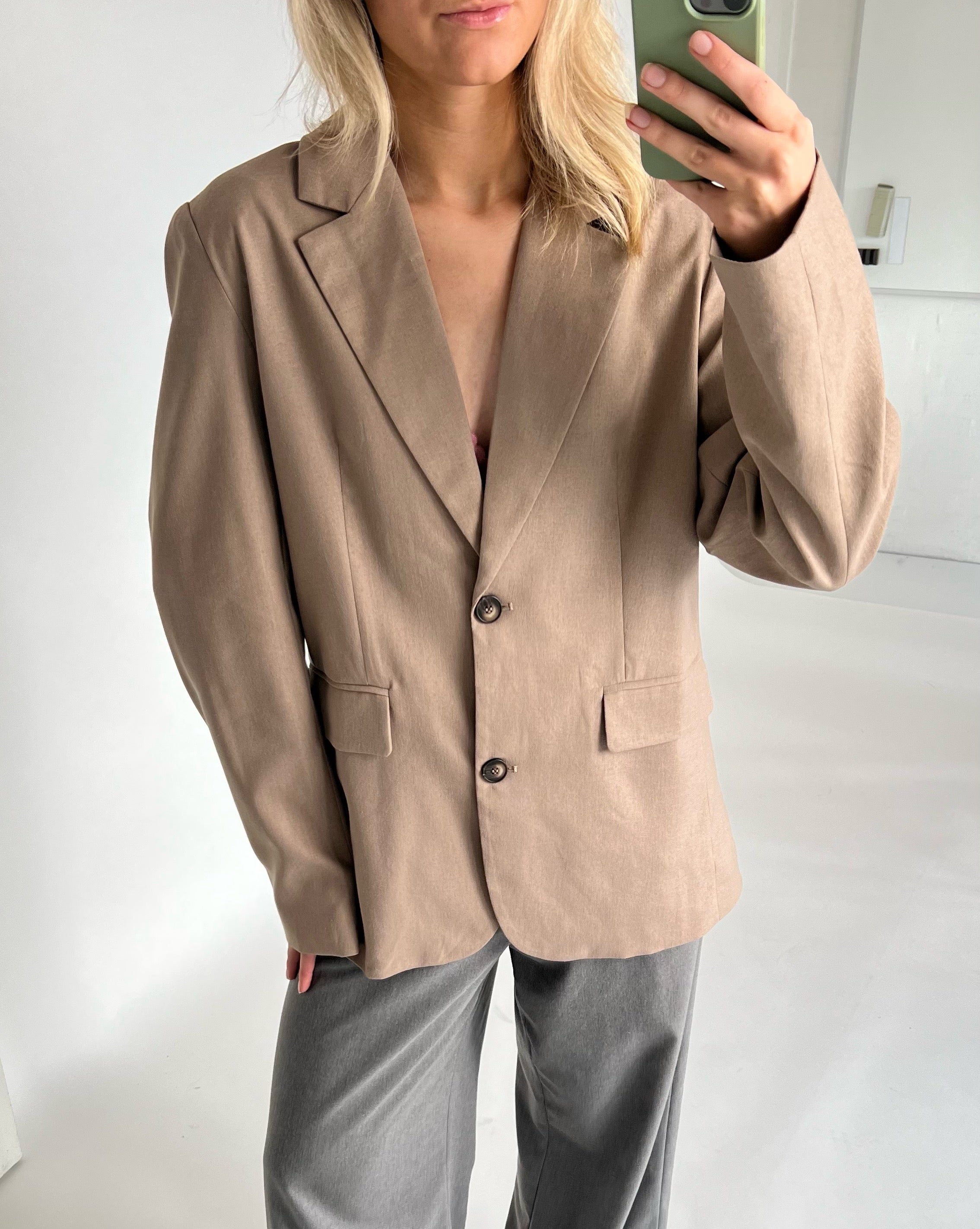 En person iført en beige blazer og grå bukser tager en selfie i et lyst rum. Personen holder en grøn mobiltelefon nær ansigtet.