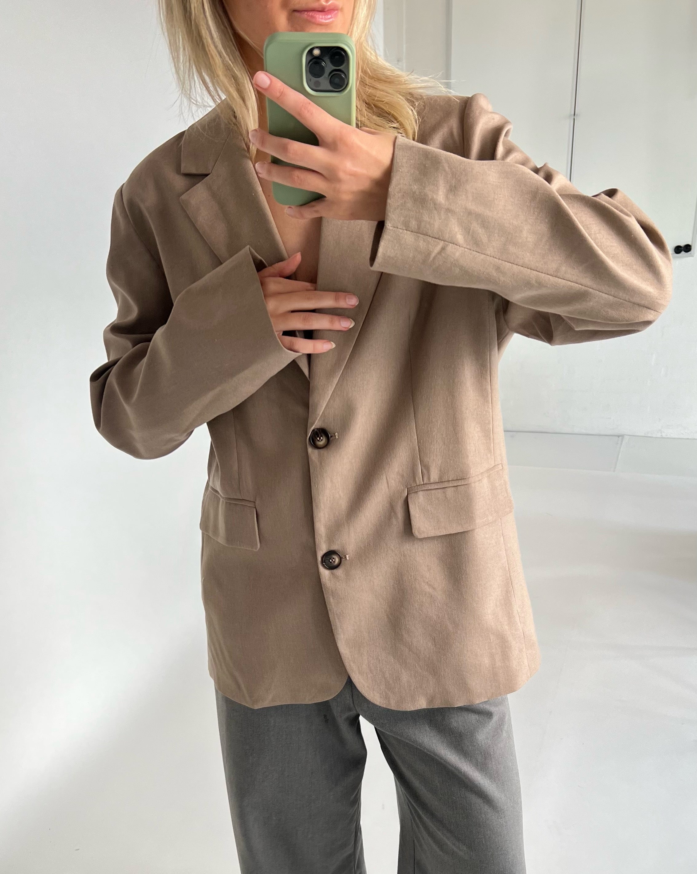 En person i brun jakke tager en selfie med en grøn telefon i et lyst rum. Personen er iført grå bukser og ses fra skuldrene og ned.