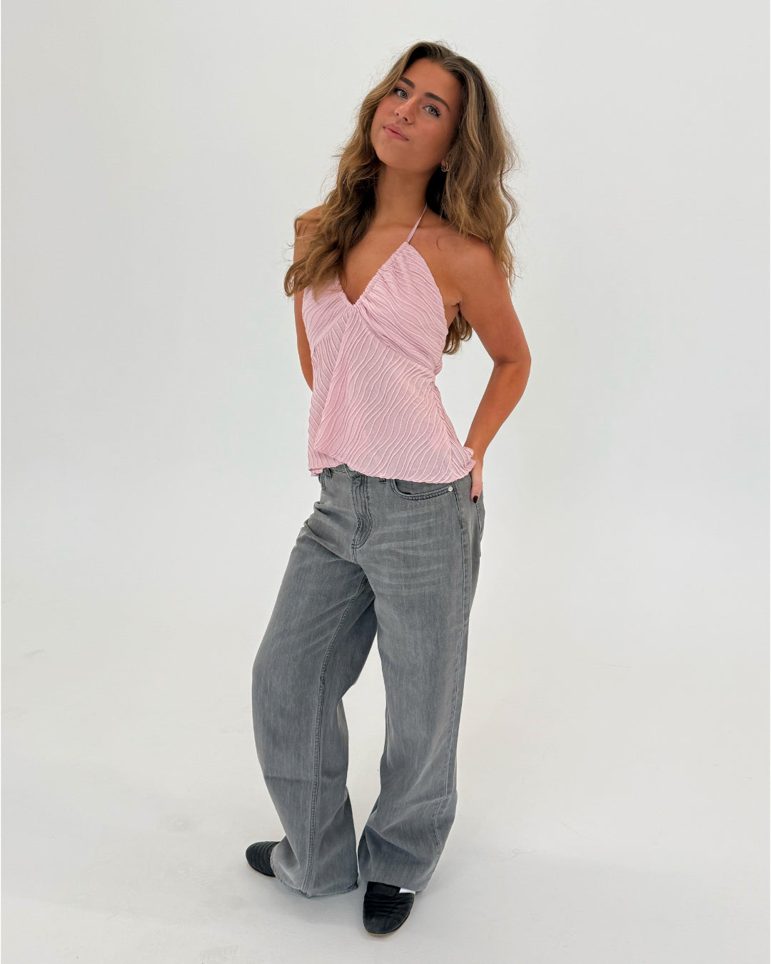 En person står i en afslappet positur iført en lyserød top og grå jeans i et rum med en neutral hvid baggrund.