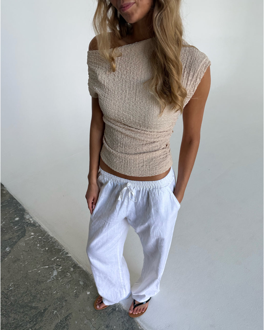 En person står iført en beige off-shoulder-top og hvide bukser med elastik i taljen. De står i et indendørs miljø med en grå betongulv og hvid væg.