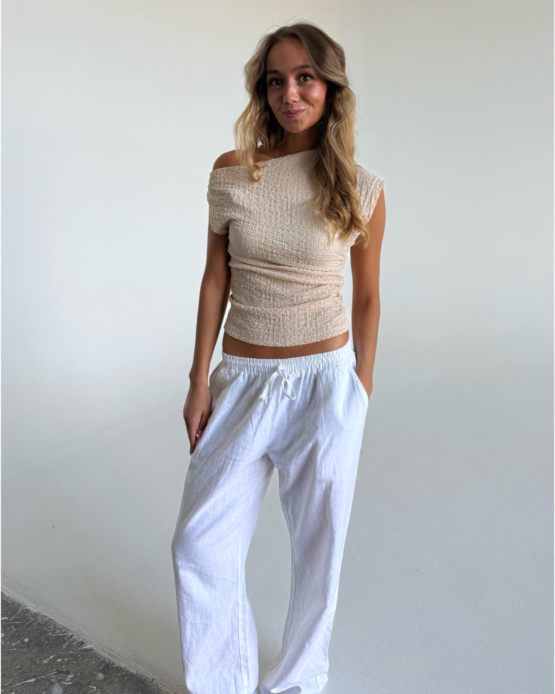 En person står afslappet iført en beige off-shoulder top og hvide bukser. Personen har hænderne i lommerne og befinder sig i et lyst, minimalistisk rum.