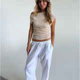 En person står afslappet iført en beige off-shoulder top og hvide bukser. Personen har hænderne i lommerne og befinder sig i et lyst, minimalistisk rum.