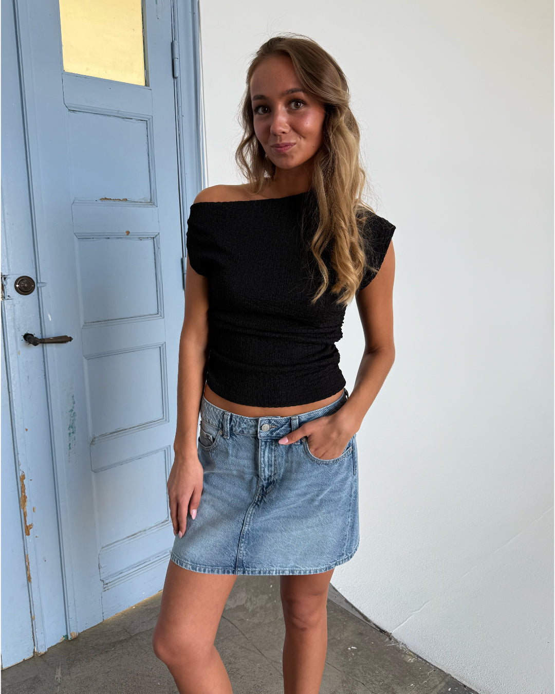 Kvinde står afslappet iført en sort top og denimnederdel. Hun har hånden i lommen. Baggrunden er en lys væg ved siden af en blå dør.