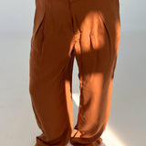 Brune bukser hænger løst på en person, der står barfodet. Sollys falder på stoffet, fremhæver folderne, i et lyst, minimalistisk rum.