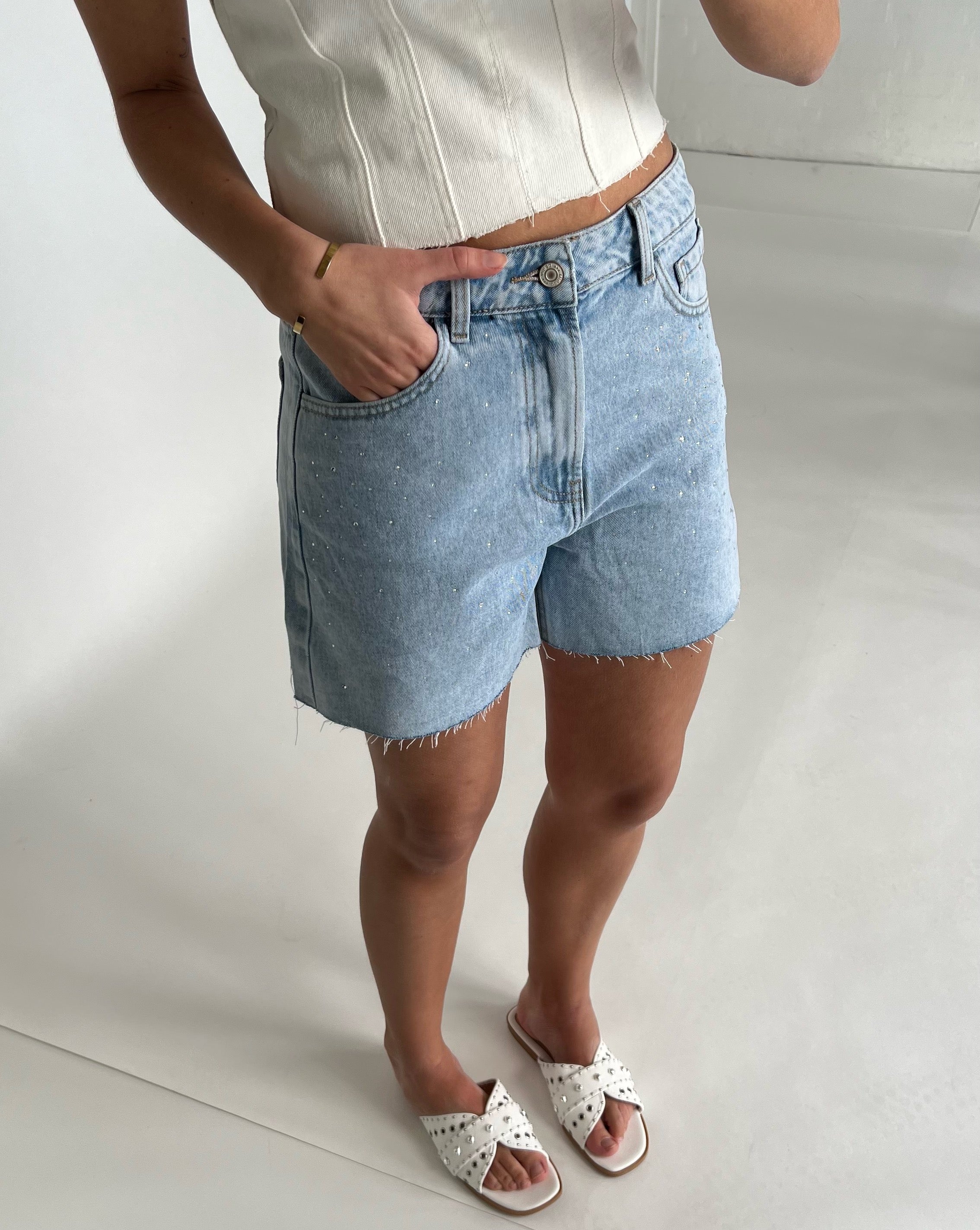 En person iført lyseblå denimshorts med en hånd i lommen står afslappet i et lyst interiør. Personen bærer en hvid top og hvide sandaler.