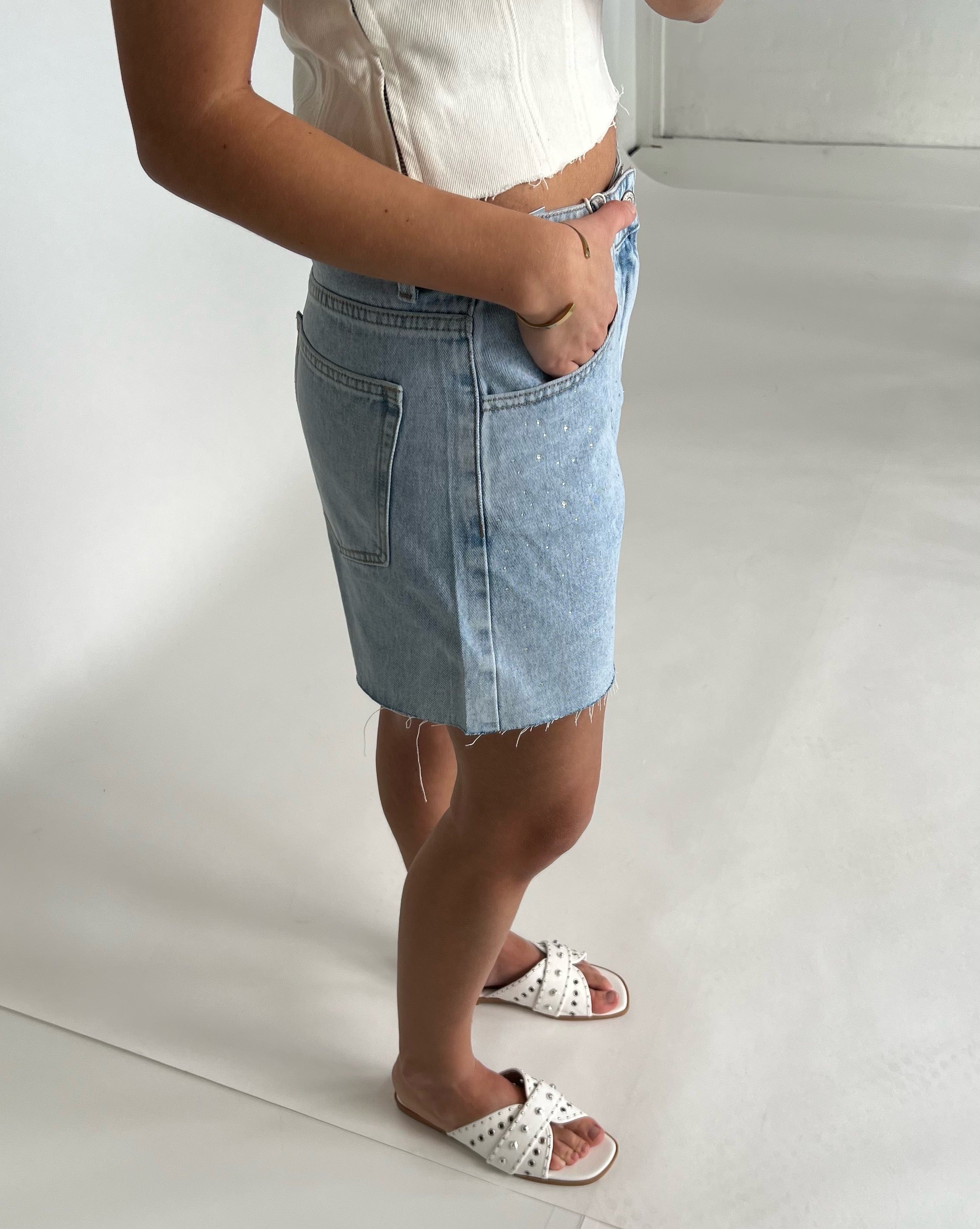En person med en hvid top og en lys denimnederdel står med hænderne i lommerne. De bærer hvide sandaler med nitter. Baggrunden er et lyst, minimalistisk rum.