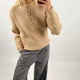 Person iført beige striktrøje med store knapper, tager et spejl-selfie med smartphone. Personen har grå bukser på og en ring på fingeren. Baggrunden er enkel og lys.