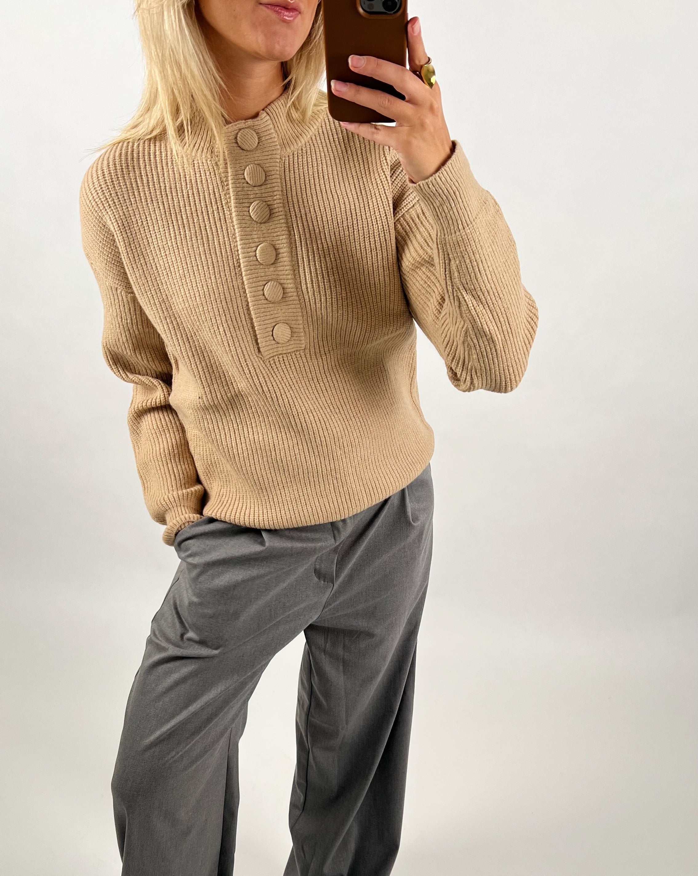 Person iført beige striktrøje med store knapper, tager et spejl-selfie med smartphone. Personen har grå bukser på og en ring på fingeren. Baggrunden er enkel og lys.