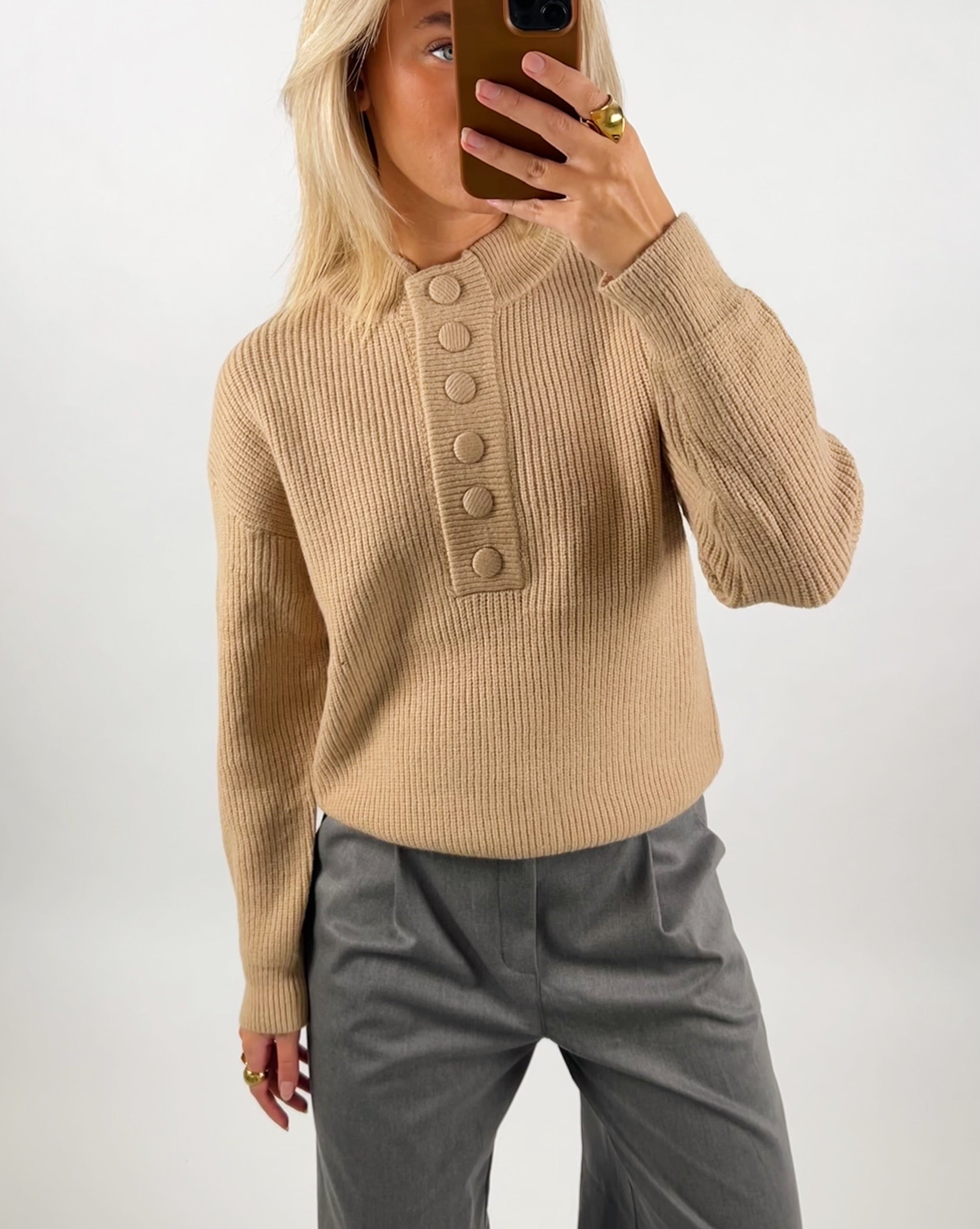 En person iført en beige striktrøje med knapper tager en selfie med en telefon. Personen har grå bukser på og bærer en ring, i et neutralt rum.