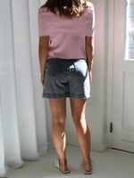 Person iført lyserød off-shoulder top og grå shorts står stille. Personen står indendørs nær et hvidt gardin og en dør, hvor sollys strømmer ind.