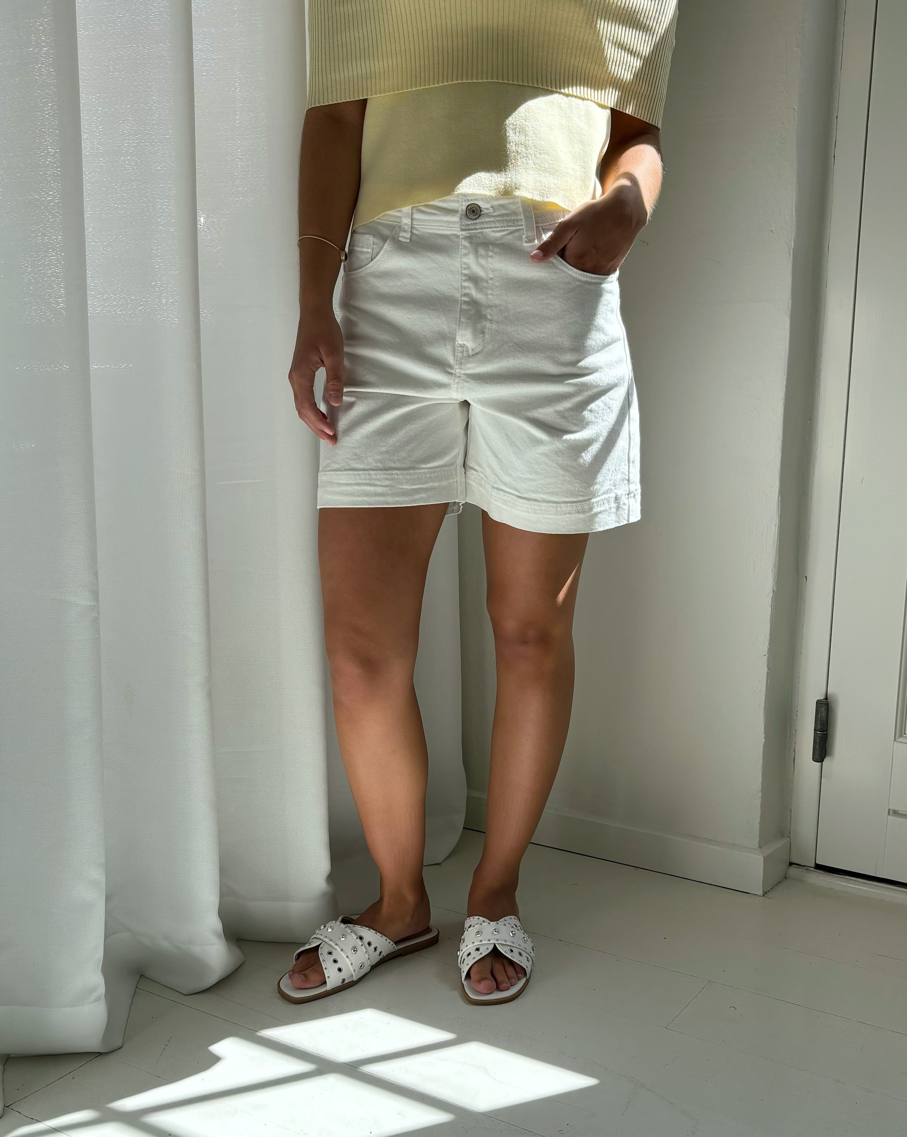 En person står iført hvide shorts og gule top, med hænder i lommerne, i et lyst rum med hvide gardiner og sollys på trægulvet.