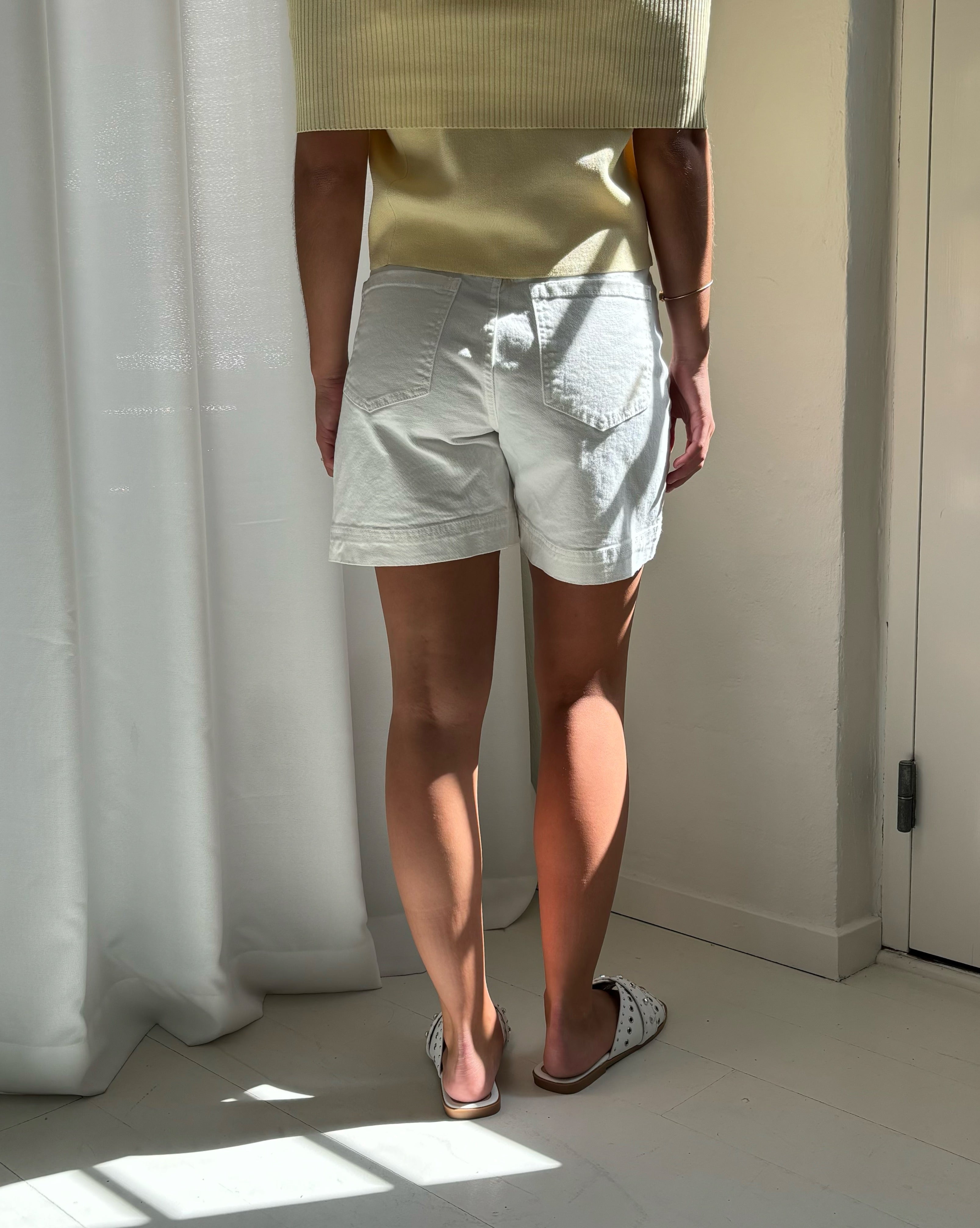 Person iført hvide shorts og gule top står stille foran hvide gardiner, badet i blødt naturligt lys i et rum med lyse vægge og gulv.