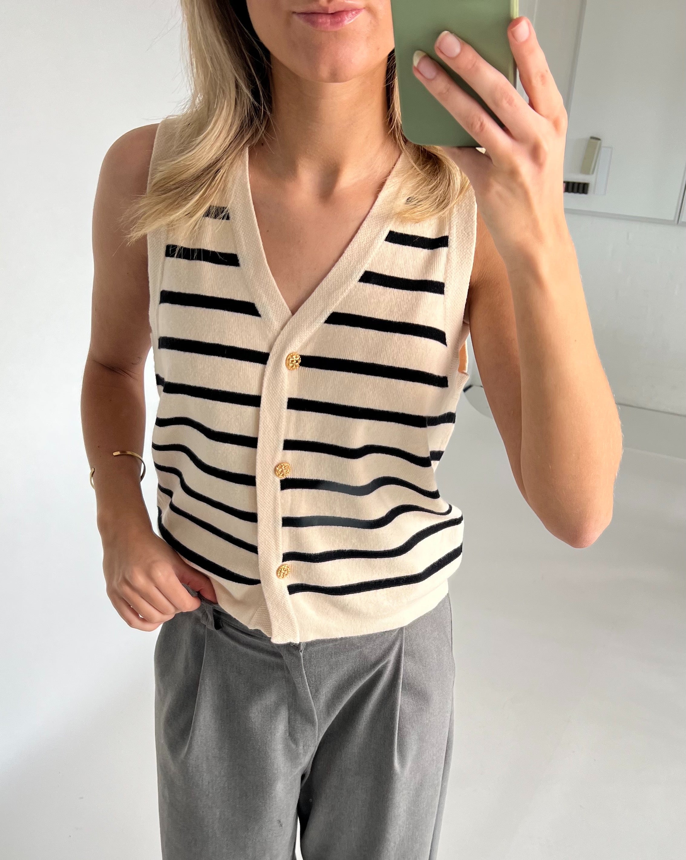 En person iført en ærmeløs, stribet vest med gyldne knapper tager et spejlbillede i et lyst, minimalistisk rum, iført grå bukser og et armbånd.