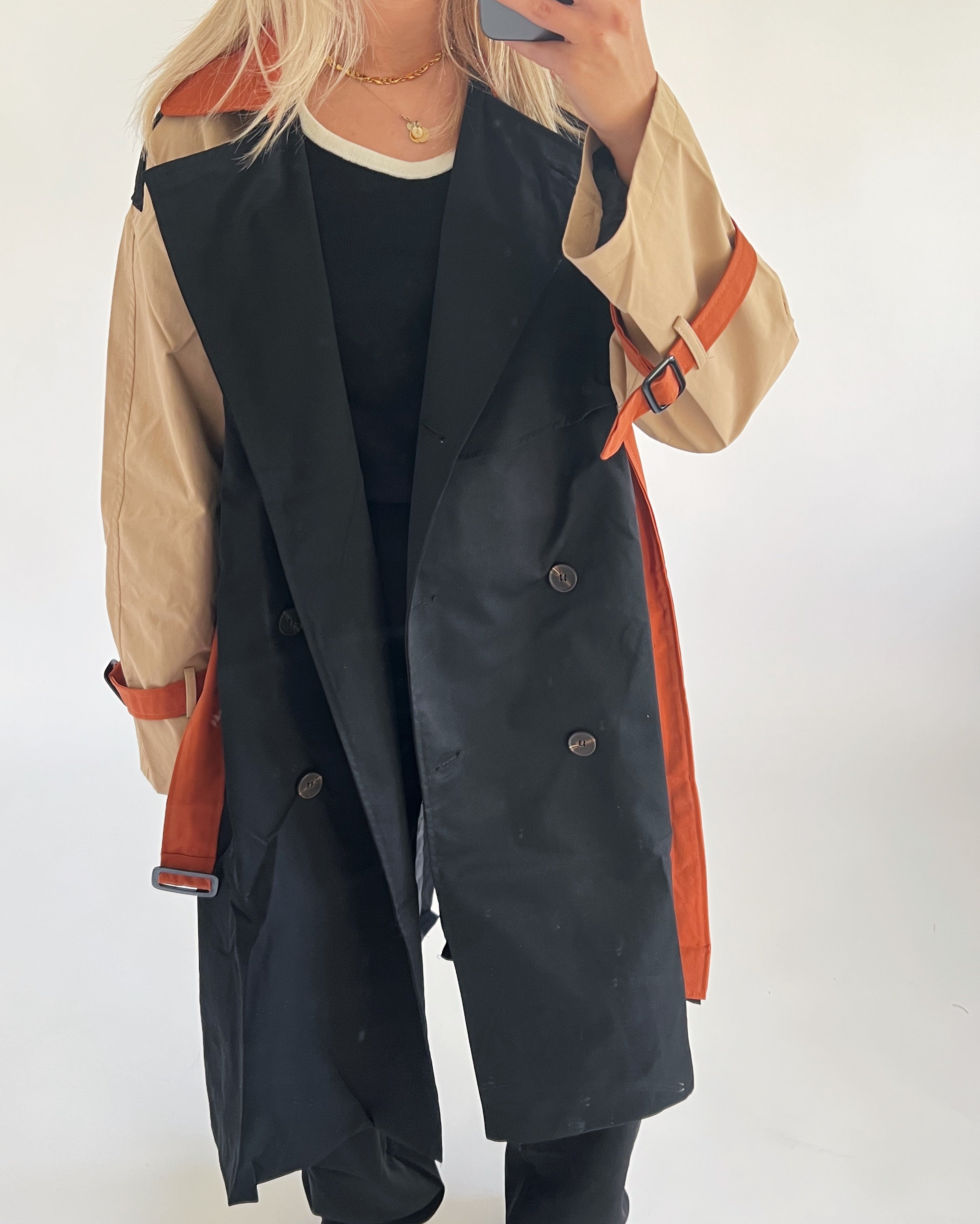 Person i sort trenchcoat med beige ærmer og orange detaljer, tager selfie. Personen står i et lyst lokale. Trenchcoaten har knapper og et bælte. Ingen tekst til stede.