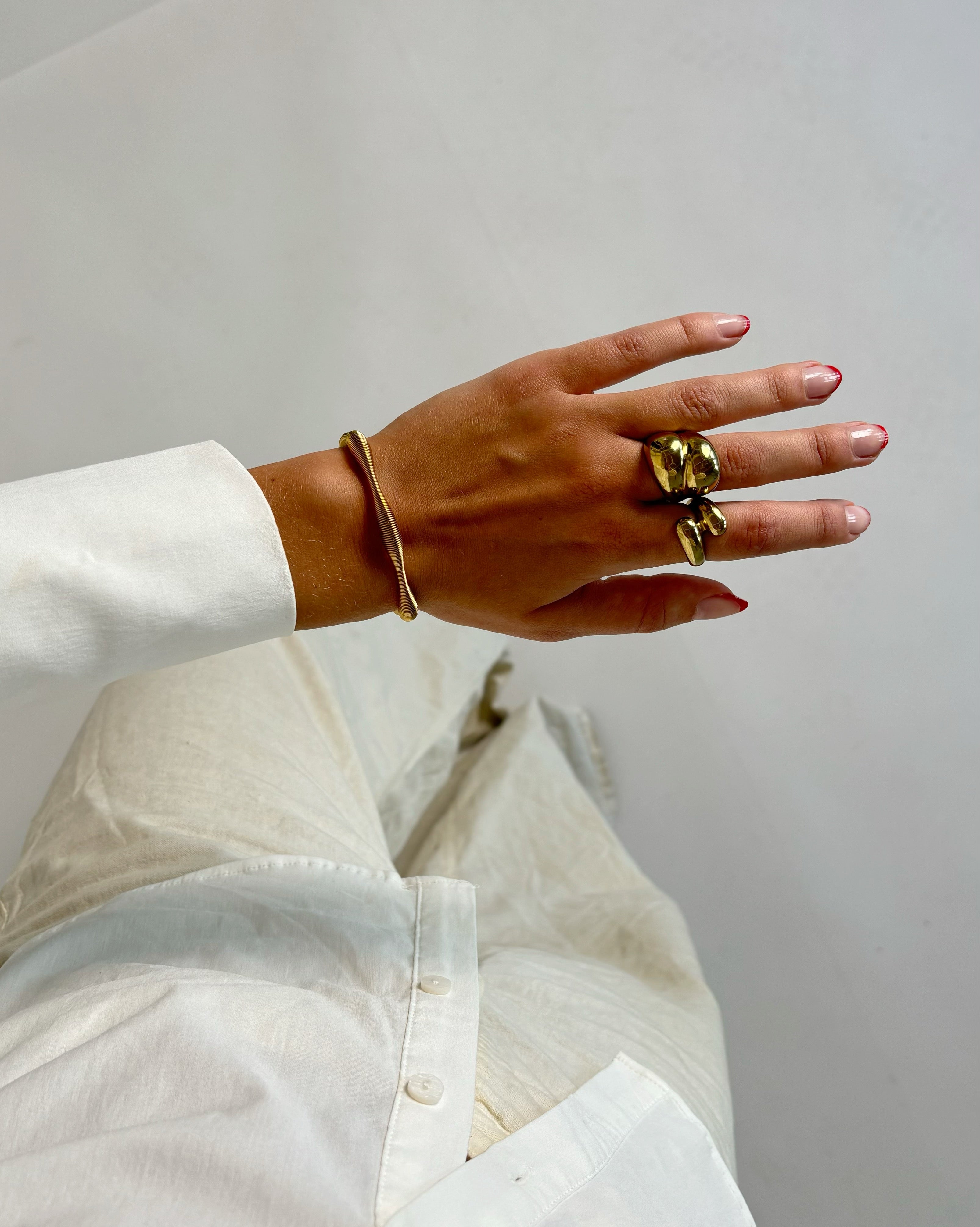 En hånd med gyldent armbånd og to skulpturelle ringe hviler over benklæder og skjorte, begge i lys stof. Baggrunden er neutral og minimalistisk.