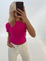 Person tager selfie med smartphone, iført lyserød bluse og hvide bukser i minimalistisk rum. Blondt hår er synligt, men ansigtet dækkes af telefonen.