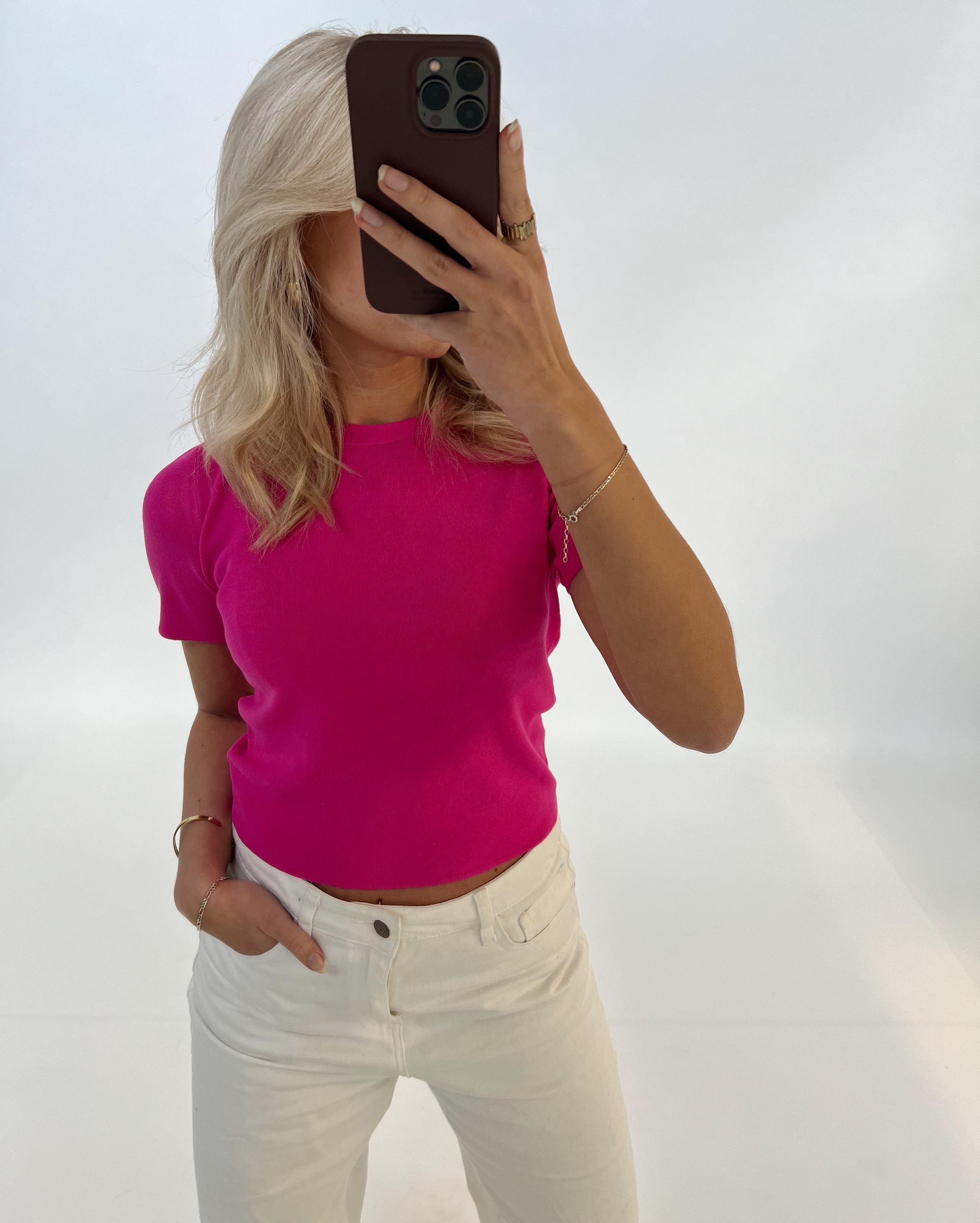 Person tager selfie med smartphone, iført lyserød bluse og hvide bukser i minimalistisk rum. Blondt hår er synligt, men ansigtet dækkes af telefonen.