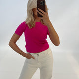 En person tager et spejlbillede, iført en pink trøje og hvide bukser, i et minimalistisk hvidt rum. Personen dækker ansigtet med en mørk telefon.