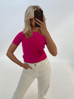 En person tager et spejlbillede, iført en pink trøje og hvide bukser, i et minimalistisk hvidt rum. Personen dækker ansigtet med en mørk telefon.
