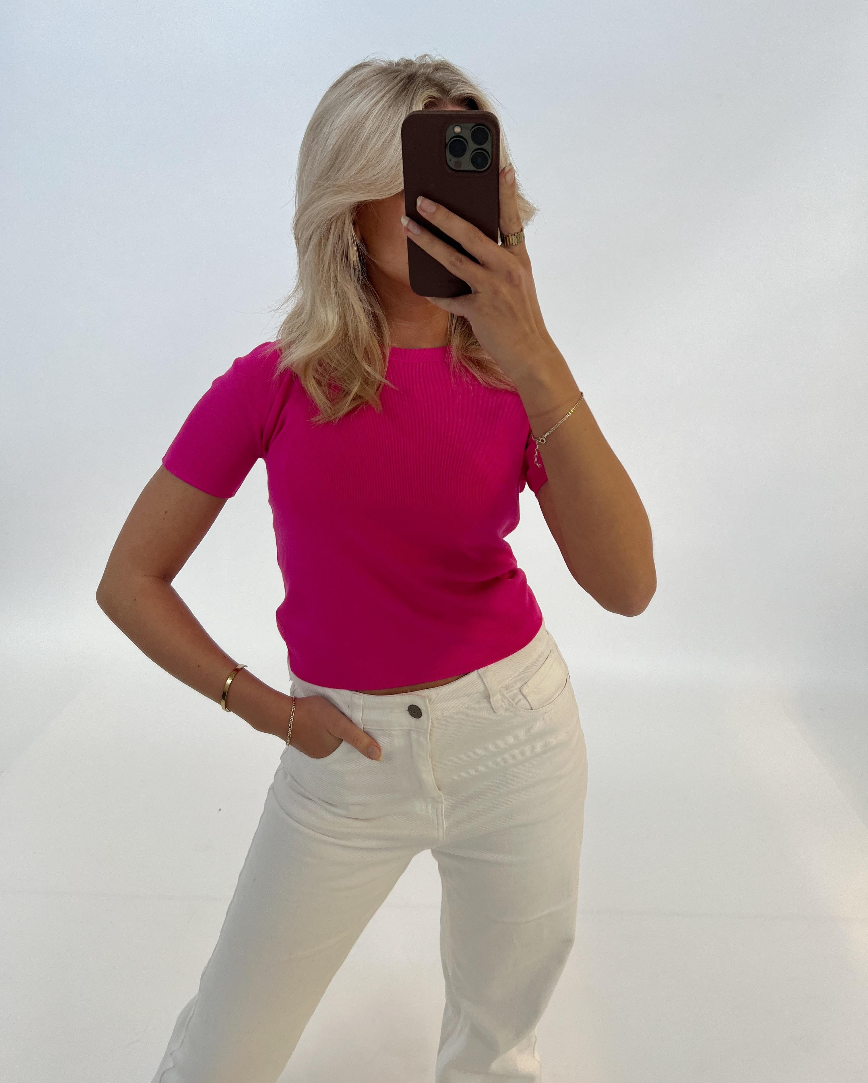 En person tager et spejlbillede, iført en pink trøje og hvide bukser, i et minimalistisk hvidt rum. Personen dækker ansigtet med en mørk telefon.