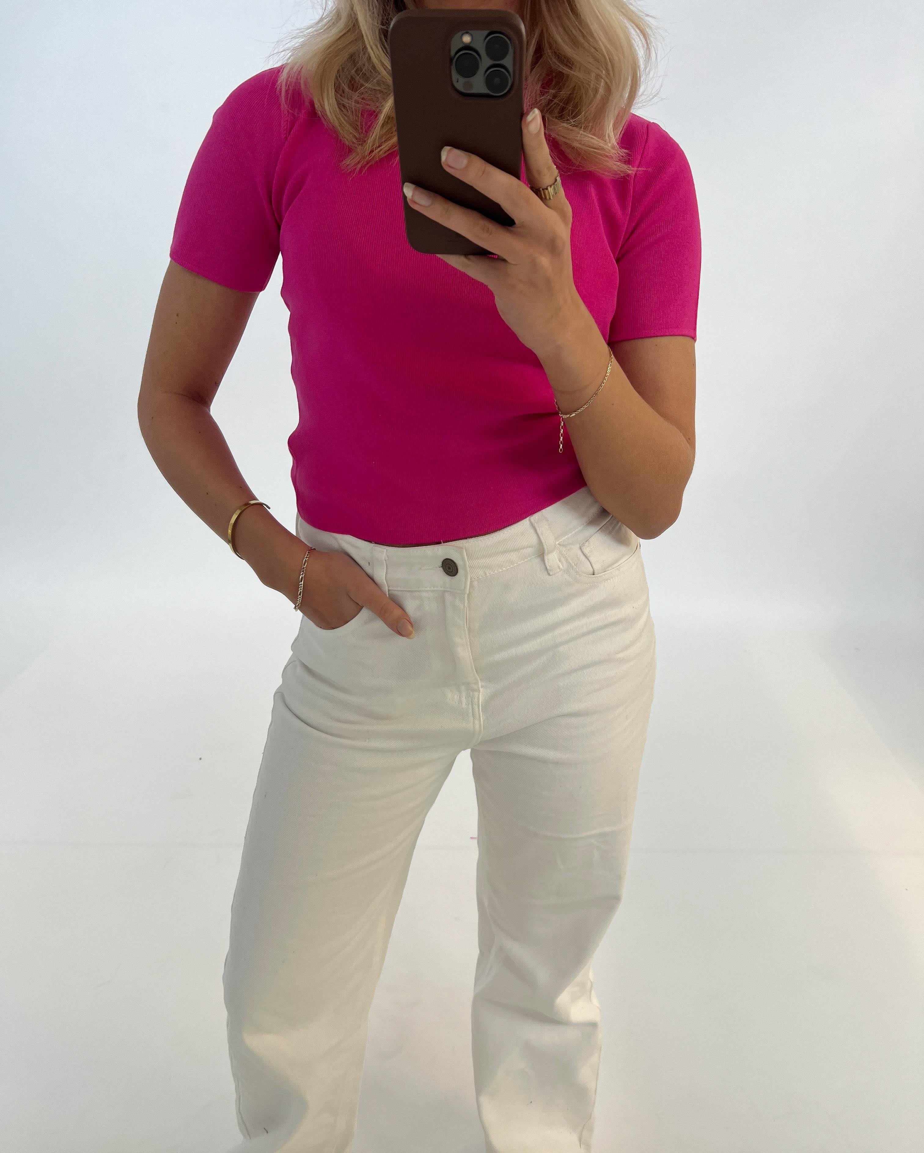 En person i pink bluse tager et billede med en smartphone, står med en hånd i lommen på hvide bukser i et hvidt rum.