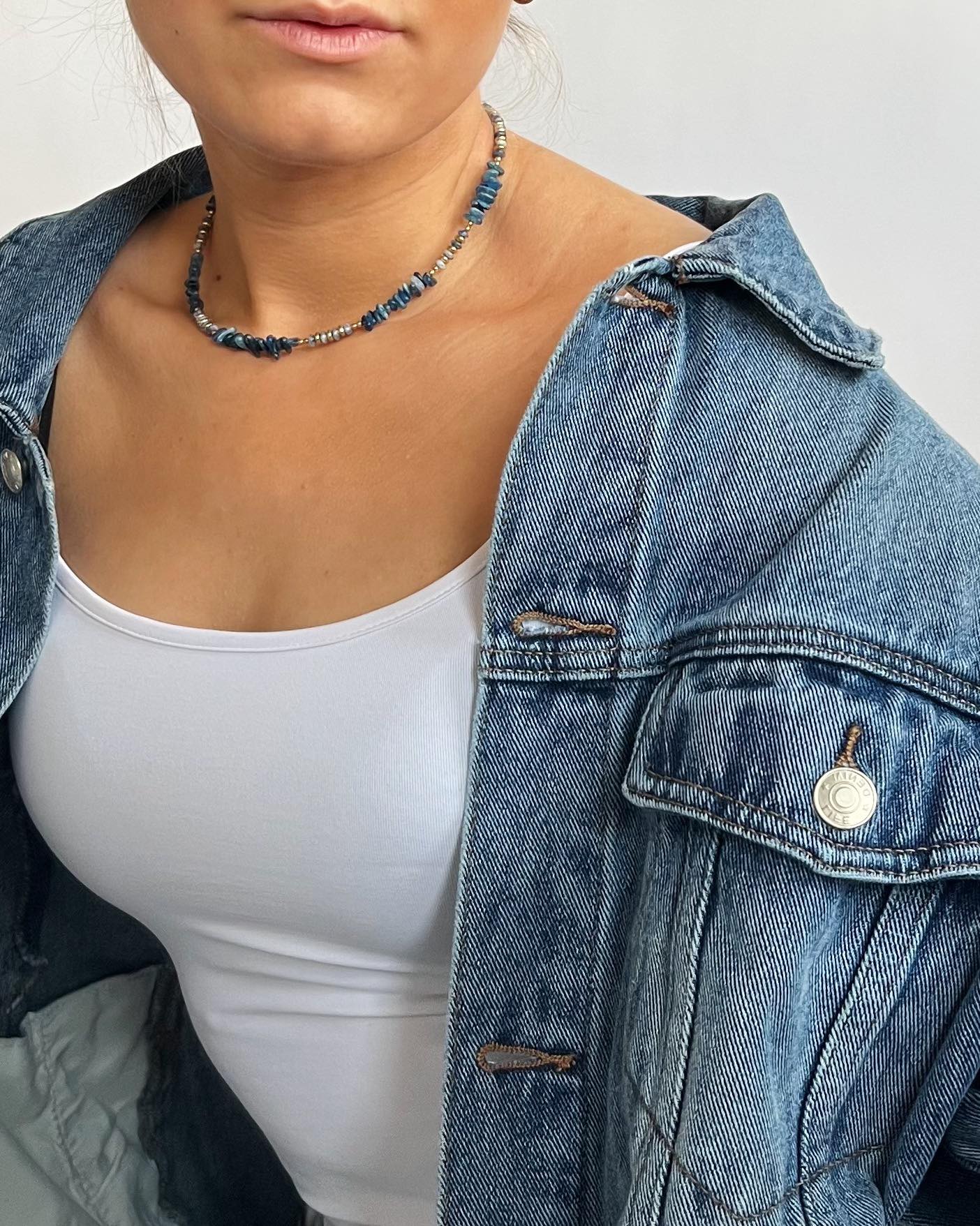En person bærer en denimjakke over en hvid tanktop og en halskæde med blå perler; personen har lys hud og står mod en lys baggrund.
