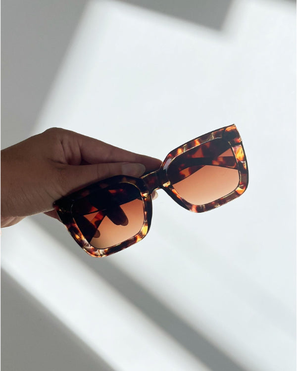 Brun, mønstret solbrille holdes i en hånd; solbrillerne har store rektangulære linser. Skygger kastes på en lys baggrund.