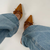 Spidse, brune sko med snørebånd og nitter bæres på fødder iført blå denimjeans. Personen står på en lys overflade.