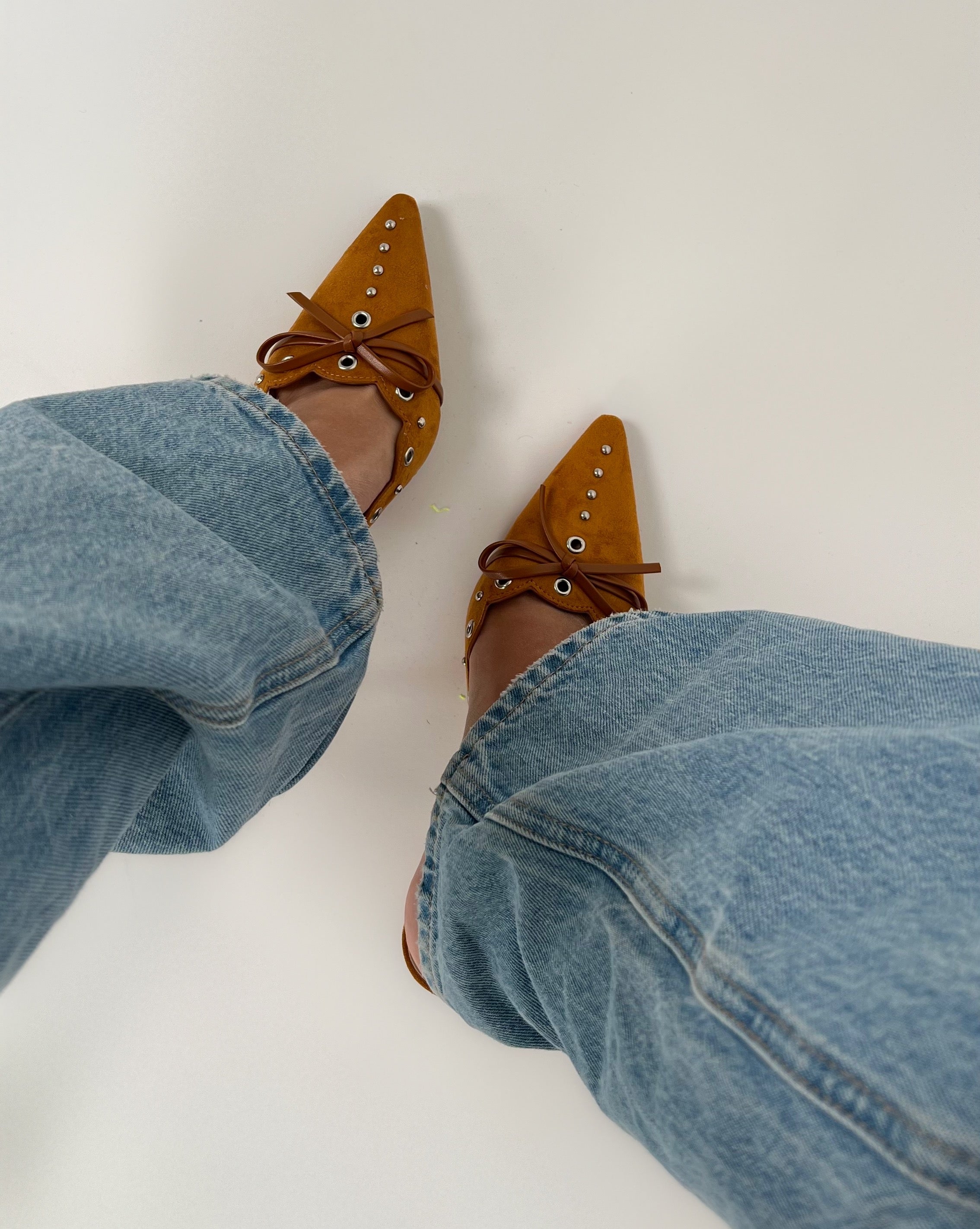 Spidse, brune sko med snørebånd og nitter bæres på fødder iført blå denimjeans. Personen står på en lys overflade.