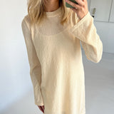 En person iført en beige sweaterkjole tager et spejlbillede med en mobiltelefon. Baggrunden er et lyst rum med hvide vægge.