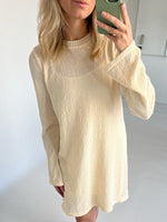 En person iført en beige sweaterkjole tager et spejlbillede med en mobiltelefon. Baggrunden er et lyst rum med hvide vægge.
