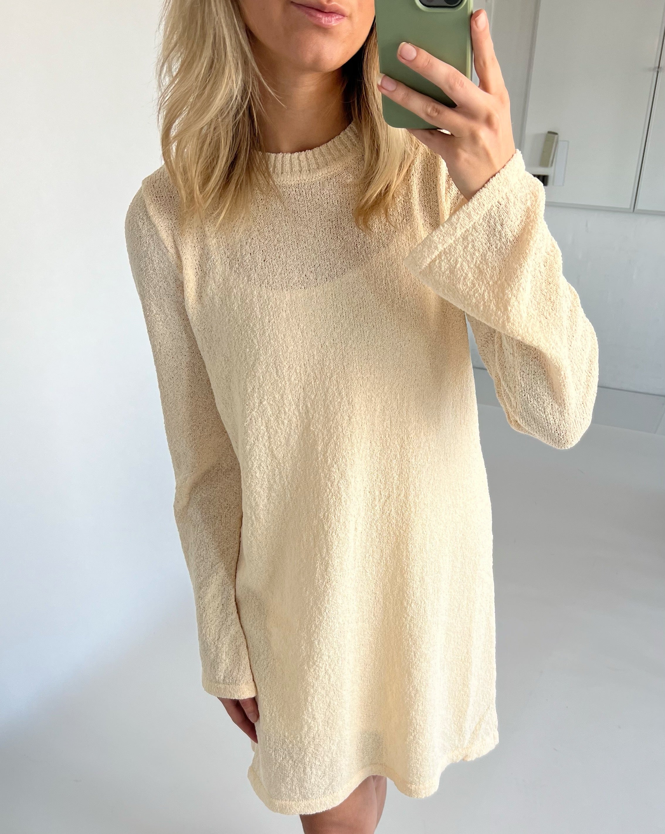 En person iført en beige sweaterkjole tager et spejlbillede med en mobiltelefon. Baggrunden er et lyst rum med hvide vægge.