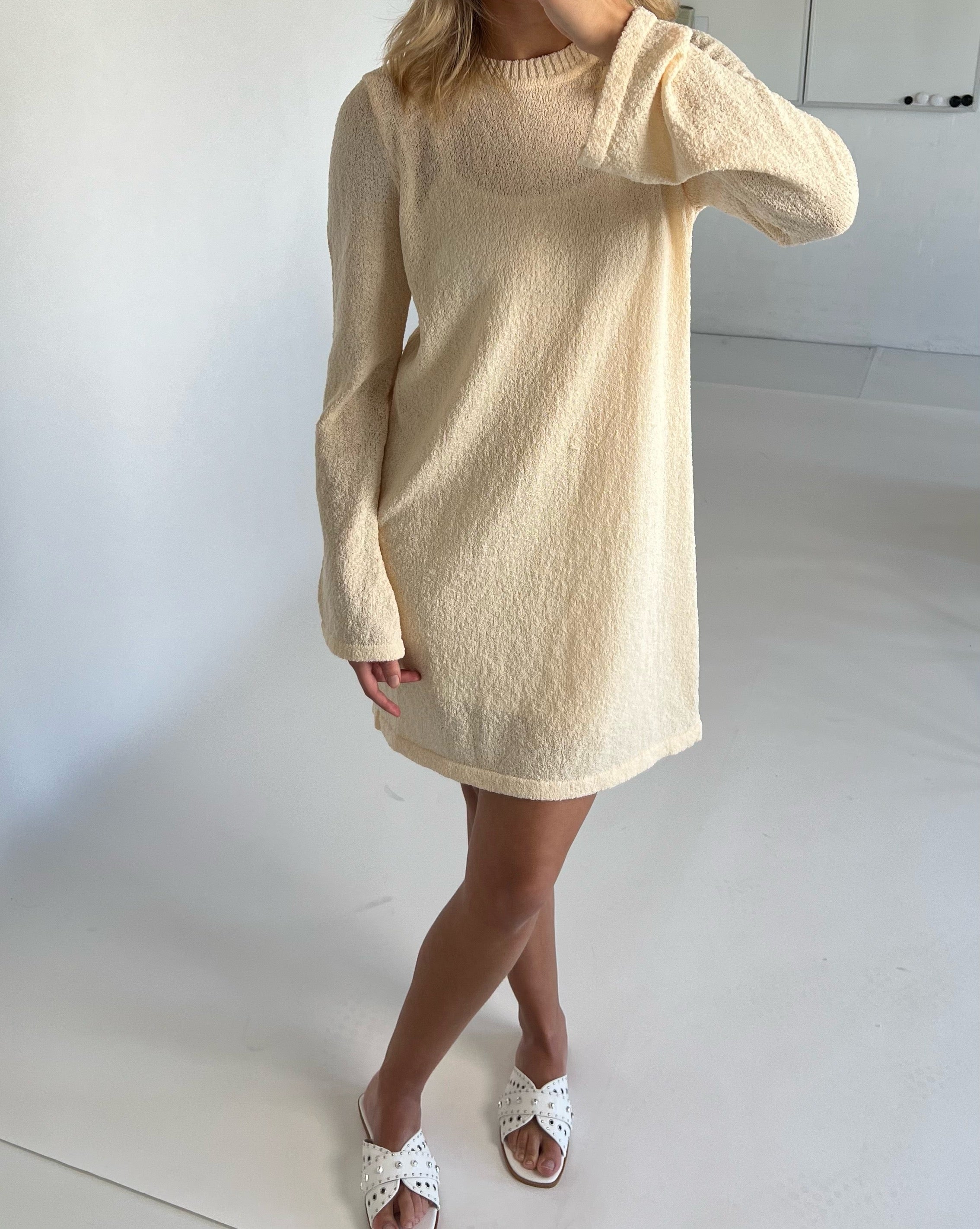En person iført en beige langærmet kjole står med krydsede ben på et hvidt gulv, iført hvide sandaler i et minimalistisk, lyst rum.