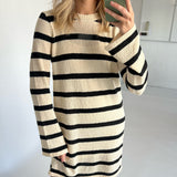 Kvinde i beige og sort-stribet kjole tager selfie med mobiltelefon i et hvidt rum.
